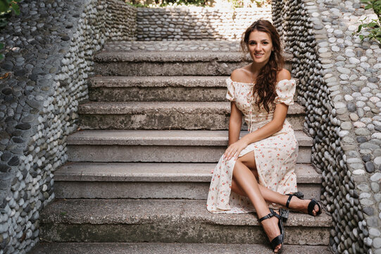 A young girl, a brunette with long hair, in a vintage dress is sitting on the steps of a gray stone staircase