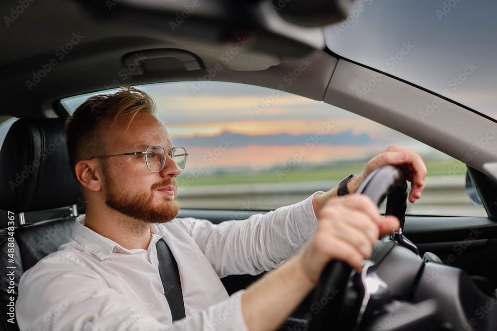 Wall mural young male driver of a car in glasses and business clothes, rides at sunset. business class driver c