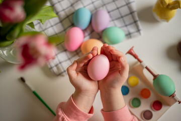 easter egg in the hands of a little girl over a table with painted eggs, watercolors and tulips.
