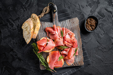 Toast with tomatoes and cured Slices of jamon serrano ham, prosciutto crudo parma on wooden board with rosemary. Black background. Top view