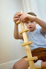 Obraz premium Cute Caucasian child diligently builds a tower of wooden bars. A boy in casual clothes plays with the details of a jenga. Educational children's games. Vertical image
