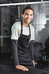 happy african american hairdresser in apron looking at camera in grooming salon.