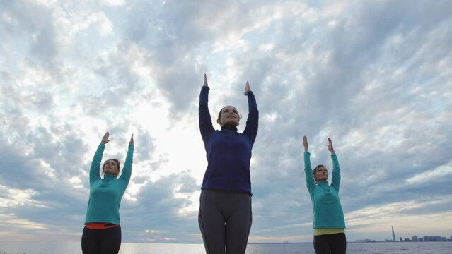 Group of three mature women practicing yoga together outdoors. Women doing standing backbend pose with raised arms standing near sea under cloudy evening sky, female instructor explaining exercise