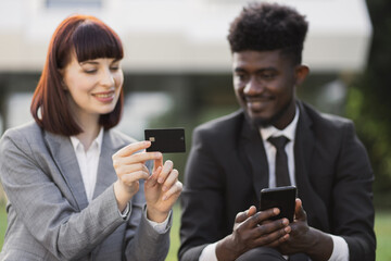 Hands of two mutlicultural business partners with bank credit plastic card and smartphone outdoors. Caucasian lady shows credit card to her male African American colleague, typing on smartphone