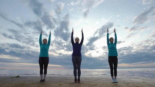 Group of three mature women practicing yoga together at sunset. Women doing standing backbend pose with raised arms near sea under cloudy evening sky, instructor teaching how to do exercise