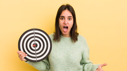 young hispanic woman amazed, shocked and astonished with an unbelievable surprise. darts target...
