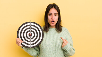 young hispanic woman looking shocked and surprised with mouth wide open, pointing to self. darts...