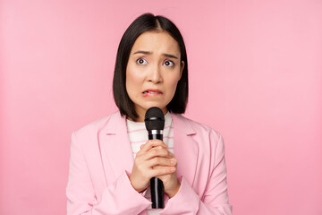 Anxious asian lady in suit, talking in public, giving speech with microphone on conference, looking...