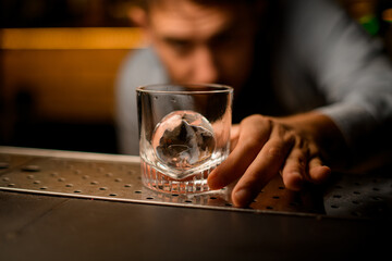 Selective focus of clean transparent old-fashioned glass with piece of ice on bar counter which male hand holds