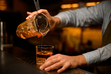 male bartender hand gently holding mixing cup with cocktail and strainer and pours bright beverage into glass