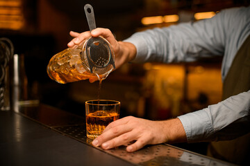 male hand gently holding mixing cup with cocktail and strainer and pours bright beverage into glass