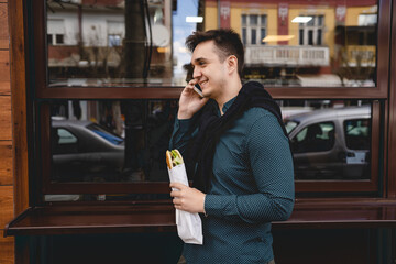 One man young caucasian male adult using mobile phone to make a call talking while holding and eating sandwich outdoor beside the building in bright day real people fast food copy space