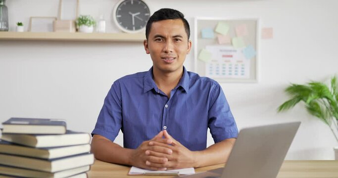 Portrait of young Asian man in blue shirt smiling and looking at camera while sitting at workdesk.