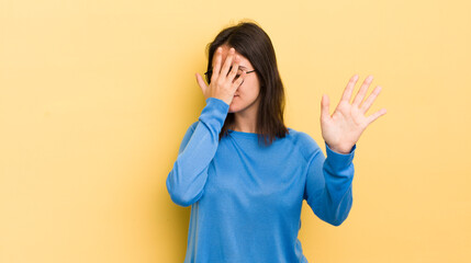 young hispanic woman covering face with hand and putting other hand up front to stop camera,...