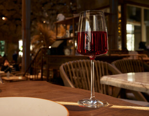 A glass of red wine on a restaurant table with illumination reflected in the glass and a blurred background.