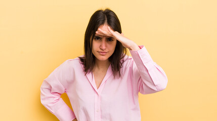 young hispanic woman looking bewildered and astonished, with hand over forehead looking far away,...