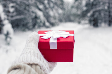 A girl in a white sweater holds a red gift box in white mitten in her hand.