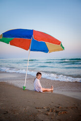 European child sits on the beach by the sea under a rainbow umbrella from the sun