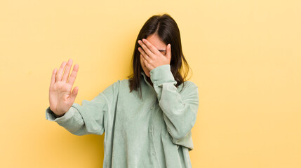 young hispanic woman covering face with hand and putting other hand up front to stop camera,...