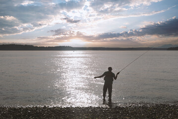 Man in silhouette casting a fly fishing pole line out over sunset lake.
