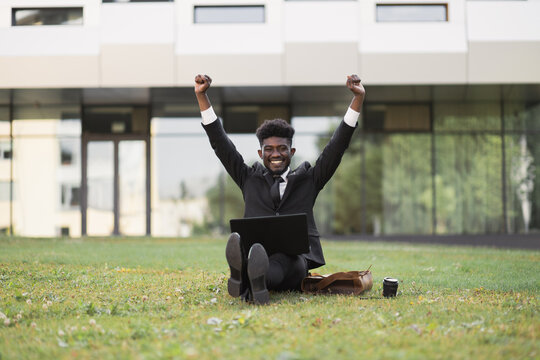 Office Work And Mindfullness. Handsome African Young Business Man With Raised Hands Working Outside In Front Of Company Building, Sitting On The Grass With Laptop And Coffee Cup. Well Done, Good News