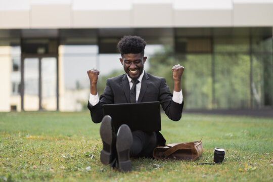 Office Work And Mindfullness. Handsome African Young Business Man With Raised Hands Working Outside In Front Of Company Building, Sitting On The Grass With Laptop And Coffee Cup. Well Done, Good News