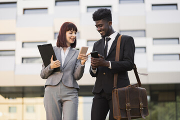 Front view of two happy smiling business colleagues, walking outside office and using smartphones. Pretty Caucasian woman and African American man in formal wear using phones outdoors