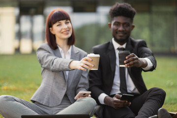 Close up serious businessman and businesswoman drinking take away coffee sitting on the grass outdoors. Portrait of business man and woman taking break with coffee to go outdoor.