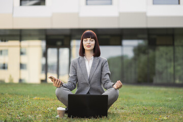 Pretty young female executive practicing yoga, sitting with eyes closed outside office on green grass, in lotus position with hands in mudra gesture, enjoying mental emotional balance during break