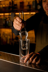 hand of bartender gently holds a piece of ice with tongs and puts it into glass