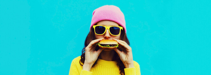 Portrait of stylish young woman eating a burger fast food on blue colorful background
