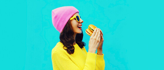 Portrait of stylish happy smiling young woman eating a tasty big burger fast food on blue colorful...