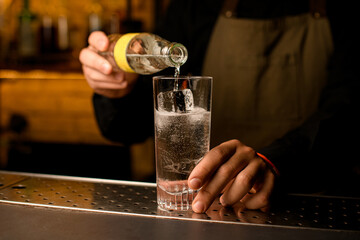 Close-up of glass with cold beverage and ice cubes into which bartender accurate pours drink from bottle