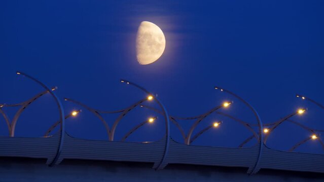 Bright moon, through a transparent veil of barely visible clouds, shine over modern high speed road. Time lapse shot at late evening, dark blue sky become darker. Cars racing by elevated highway