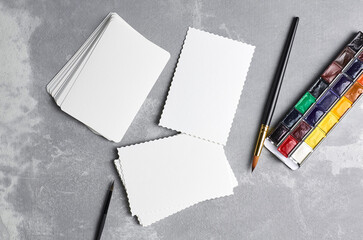 Flat lay with blank paper cards, watercolor palette and paint brush on grey concrete baclground. Mockup for artwork presentation, top view, copy space