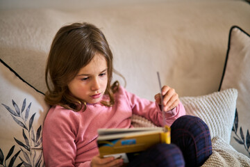 A little girl sitting on a couch and reading a book.