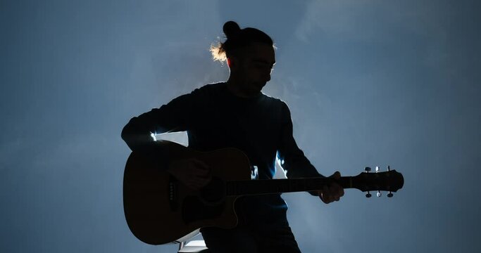 Silhouette of young man playing guitar sitting on stool in illuminated background and smoke