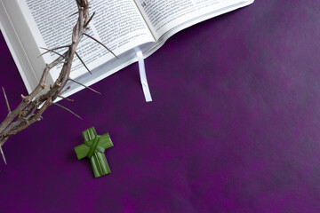 Palm leaf cross and crown of thorns on open bible on a dark purple background with copy space