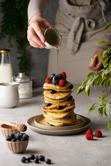 Female pouring honey maple syrup on traditional blueberry pancakes with raspberries and walnuts stacked on top of each other