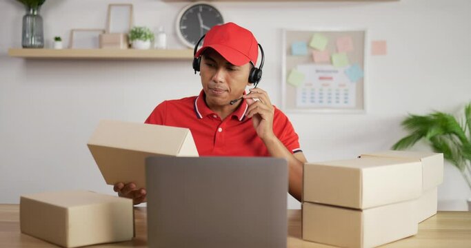 Portrait of Young Asian busy postman in red uniform, headphone and cap sitting at desk and talking to customer in postal office store and looking at clipboard. Parcels front side.
