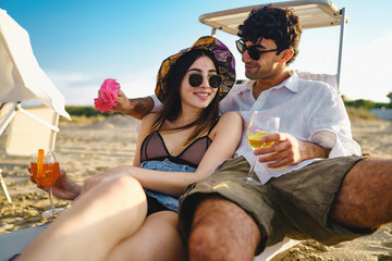 Romantic couple on vacation in the summer sitting on the chaise longue on the beach drinking fruit...