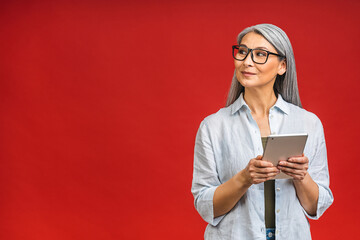 Portrait of smiling beautiful asian senior aged mature woman using tablet computer, isolated over red background. Copy space for text.