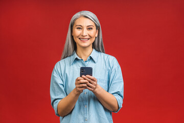 Happy mature senior asian woman holding smartphone using mobile online apps, smiling old middle aged lady texting sms message chatting on phone isolated over red background.