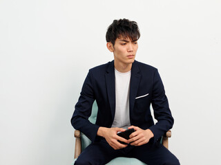 Portrait of handsome Chinese young man in dark blue leisure suit sitting in armchair and posing against white wall background. Legs crossed with mobile phone in hand, looking away and thinking.