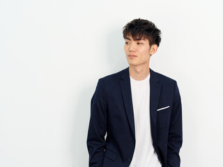 Portrait of handsome Chinese young man in dark blue leisure suit standing and posing against white wall background. Smiling and looking away, front view.