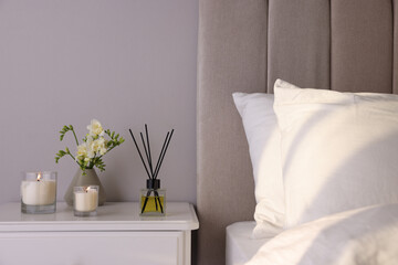 Aromatic reed air freshener, freesia flowers and candles on white bedside table in bedroom