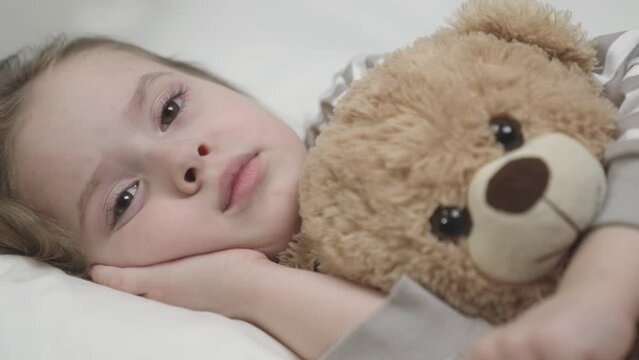 Sleepy kid girl lies bed soft pillow hugging bear toy. Sleepy melancholic small girl lies bed soft pillow hugging chest teddy bear toy. Tired child puts palm under remembering past day with sadness.