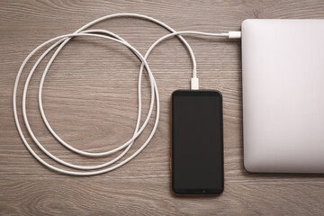 Smartphone connected with charge cable to laptop on wooden table, flat lay. Space for text
