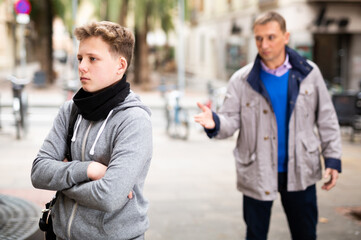 Frowning teenager listening to reprimanding from his father on city street..