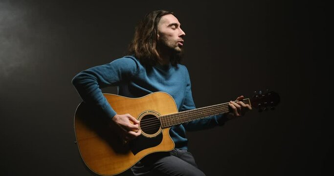 Young man singing and playing guitar sitting on stool in black background